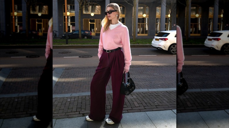A woman wearing a petal pink sweater with burgundy pants while posing on the street