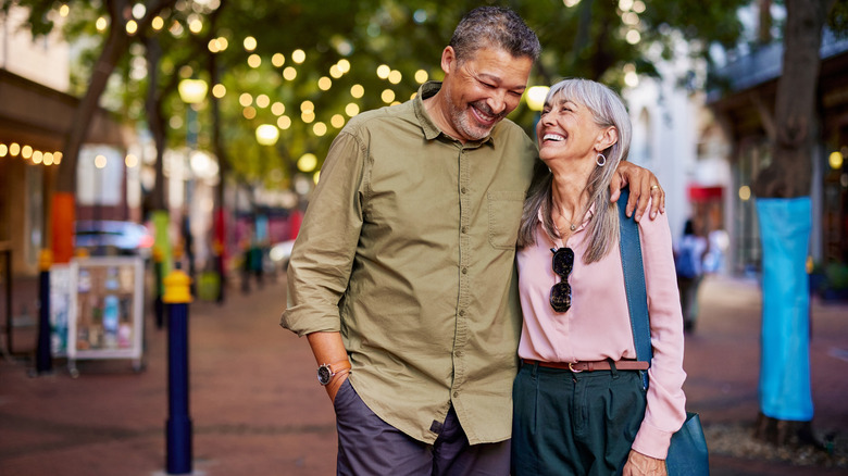 An older man and woman laughing and walking down a street with their arms around each other