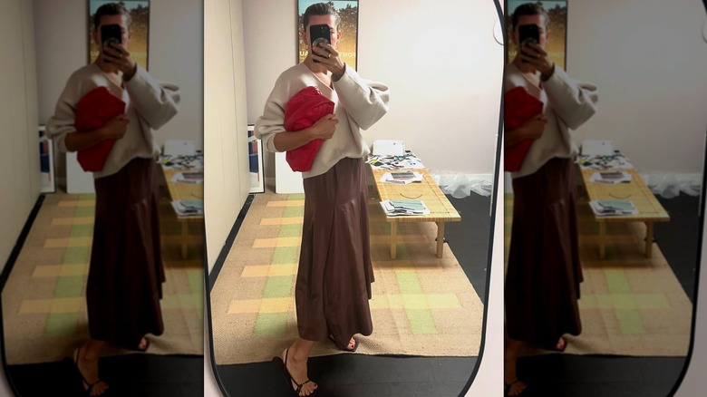A woman taking a mirror selfie of herself in a brown skirt, cream sweater, and black sandals while holding a red clutch