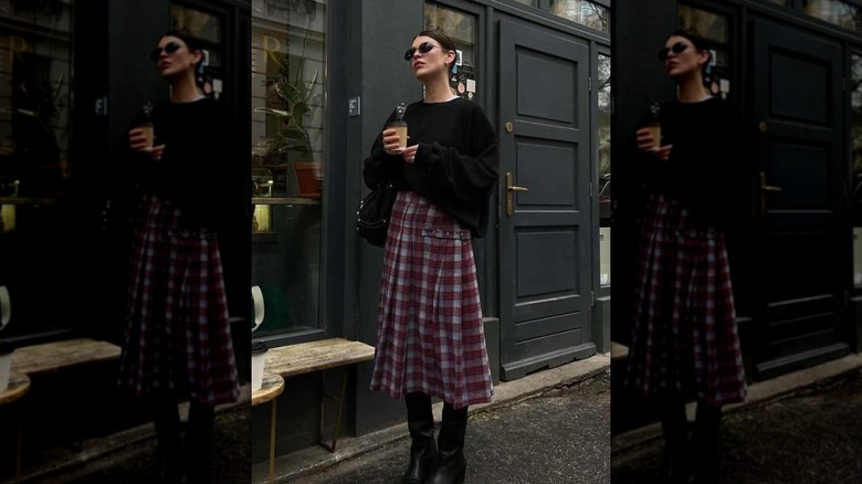 A woman standing on the street holding a cup of coffee and wearing a plaid midi skirt