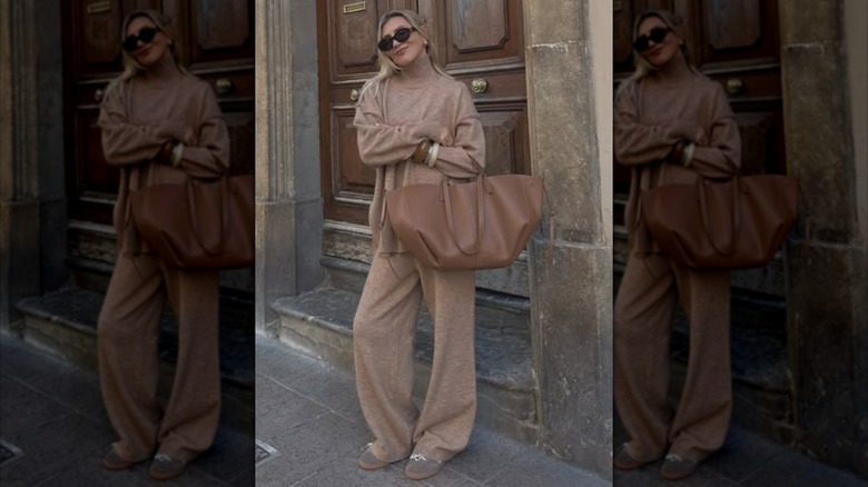 A woman standing on the street wearing a light brown knit set and holding a big leather tote