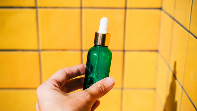 A hand holding a green serum bottle in front of a yellow tiled wall