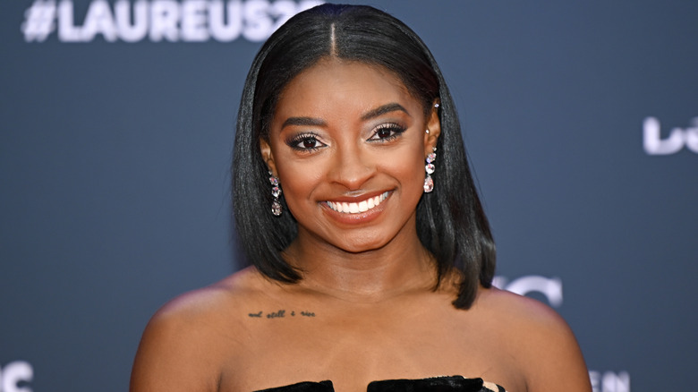 Simone Biles smiling while looking into the camera at a 2025 event, with her collarbone tattoo visible