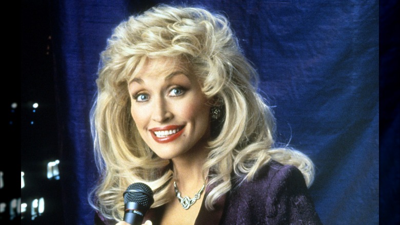 Dolly Parton smiling and holding a microphone while wearing an excessive wig in a 1992 promo photo