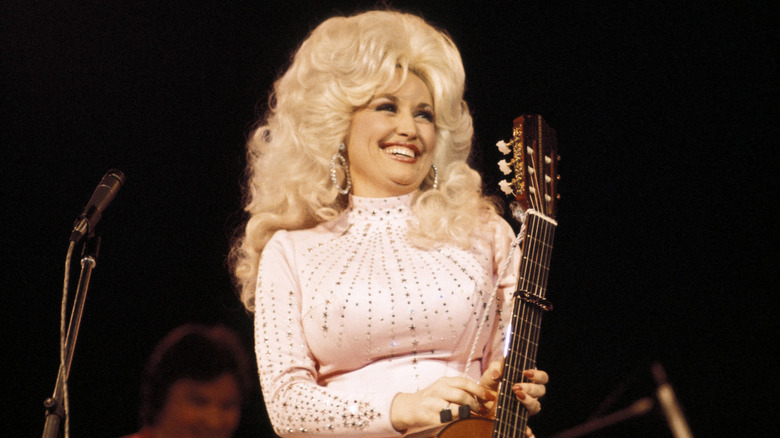Young Dolly Parton laughing onstage while wearing a white costume and tall, curly wig