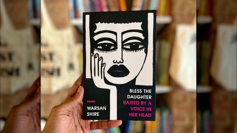 A hand holding a copy of "Bless the Daughter Raised by a Voice in Her Head" in front of a bookshelf that's out of focus