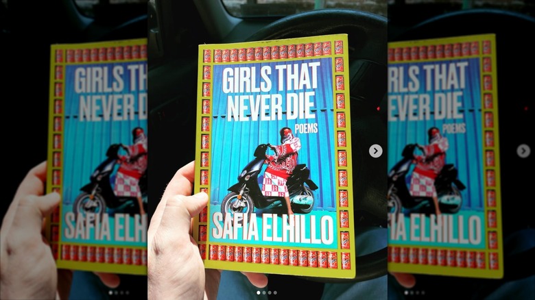 A hand holding a copy of "Girls That Never Die" in front of a steering wheel in a car