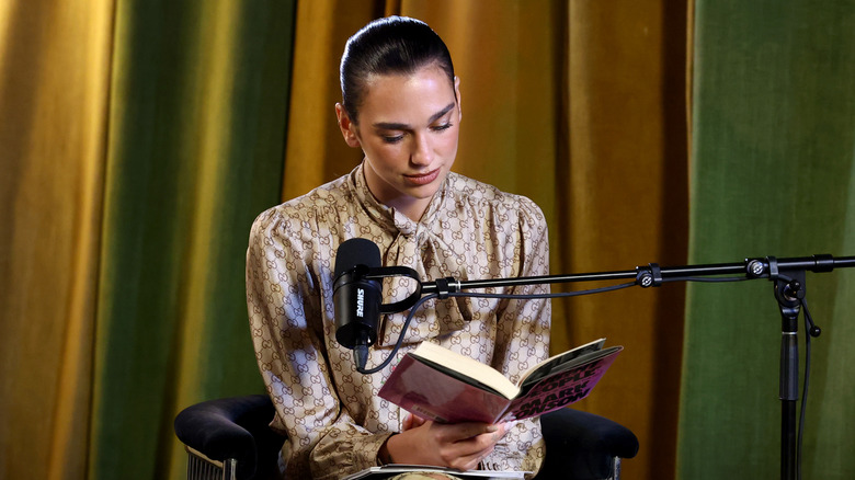 Dua Lipa sitting in a recording studio near a microphone while reading a book