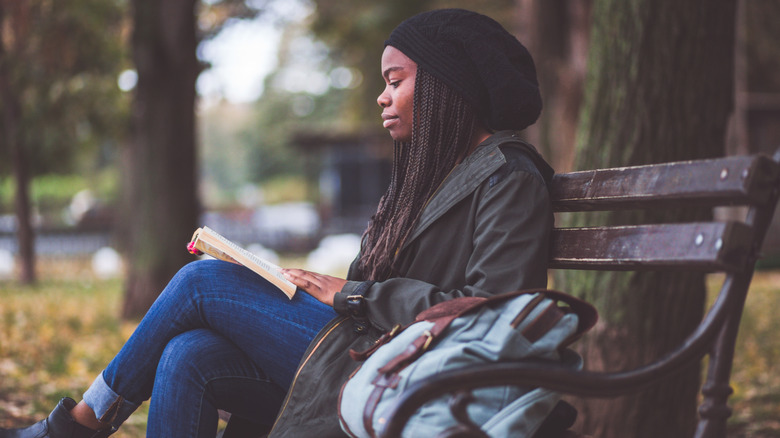 Woman with braids reading a book on a park bench during a fall day
