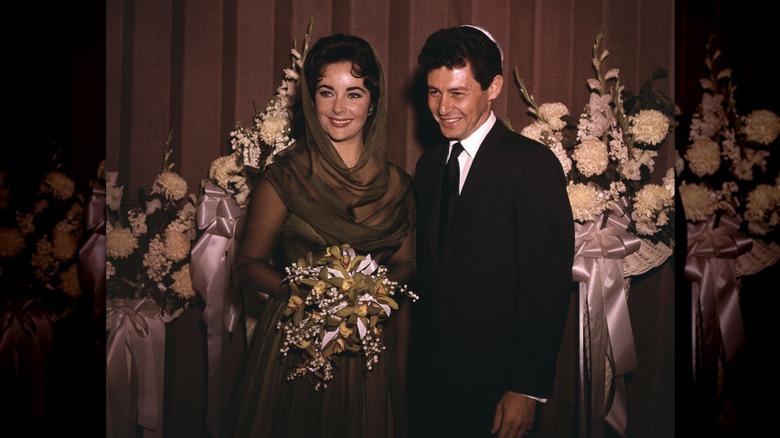 Elizabeth Taylor wearing an oliver-green dress with sheer sleeves and a matching headscarf while standing with Eddie Fisher