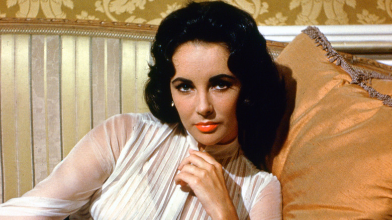 Elizabeth Taylor posing on a couch and pillow while wearing a white blouse and orange-toned lipstick
