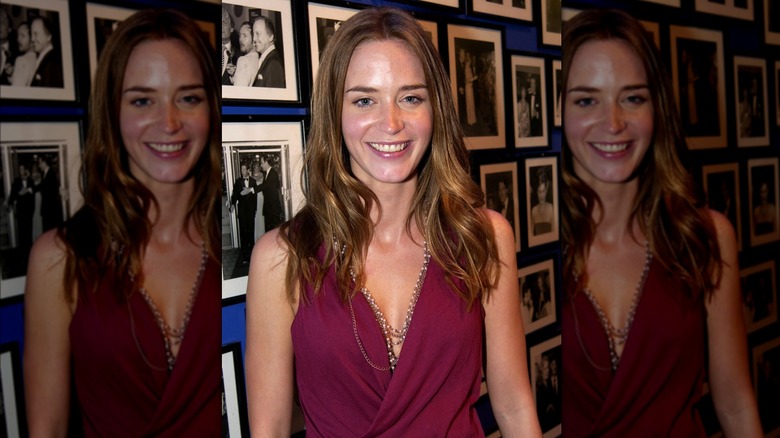 Emily Blunt smiling in front of a wall of framed photos in 2004