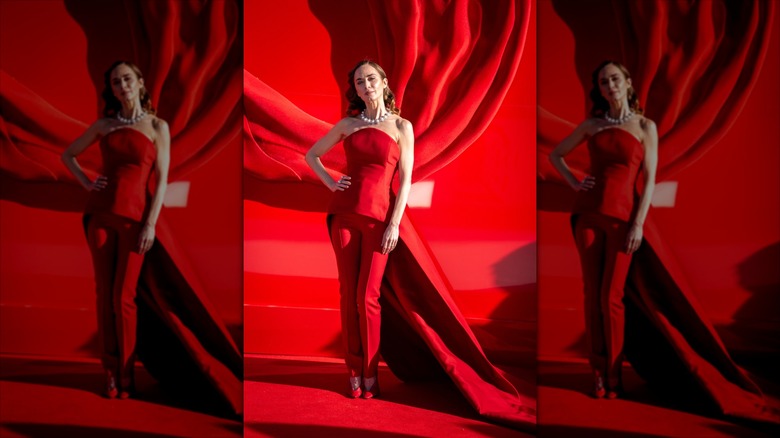 Emily Blunt poses wearing a red pantsuit and pearl choker.