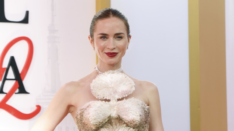 Emily Blunt smiles wearing red lipstick and a feathered top.
