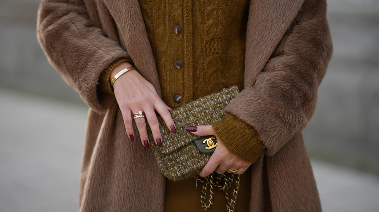 A close-up of a woman with merlot nail polish wearing a brown coat and brown sweater while carrying a purse