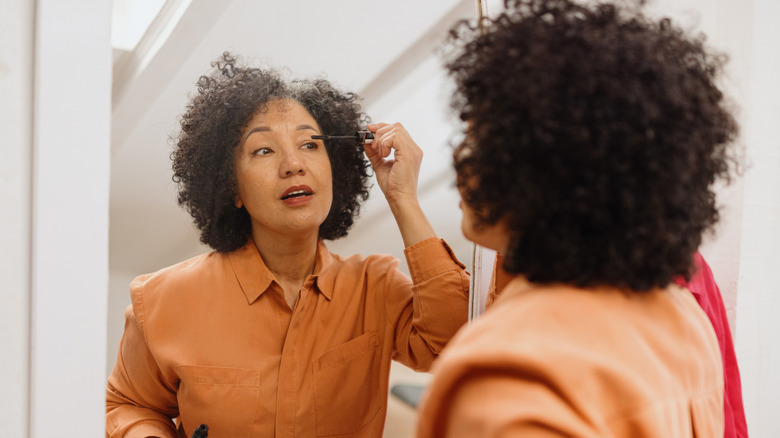 Mature, curly-haired woman applying mascara in a full-body mirror