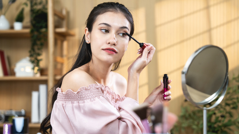 A woman applies mascara to her eyelashes while looking into a mirror