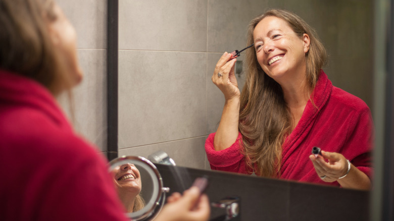 Senior woman's happy reflection seen in mirror as she applys mascara while wearing a red robe