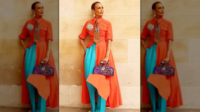A stylish older woman in an open orange dress with turquoise pants and a purple bag