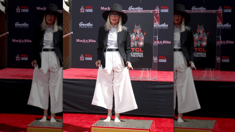 Diane Keaton stands wearing white wide-leg trousers and a black bowler hat