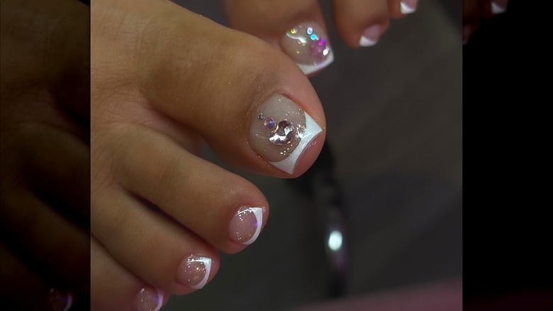 A close-up of a French pedicure with rhinestone accents and a glitter coat by b0mbnailz