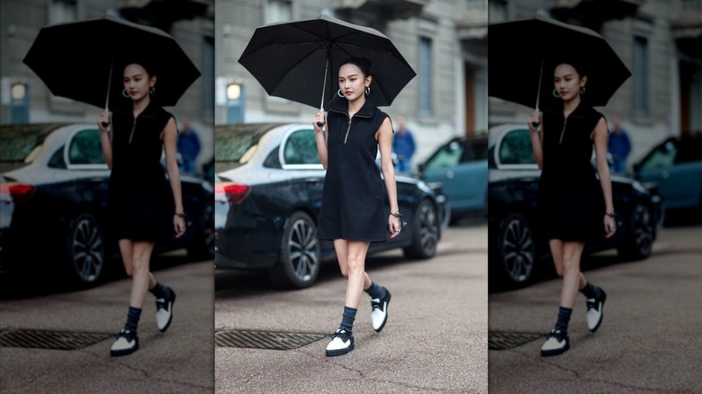 A woman walking on the street with an umbrella, black minidress, and band and white derby shoes