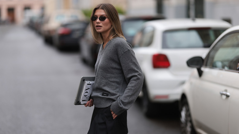A woman in a grey V-neck sweater with small sunglasses outside