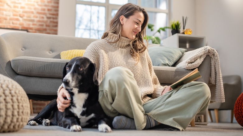 A woman sitting on the floor in a living with reading a book with a dog by her side