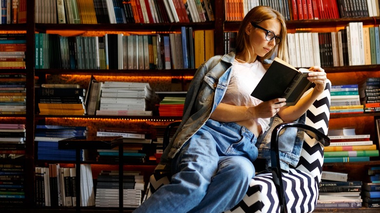 A woman sitting on a chair reading a book in front of a series of bookshelves