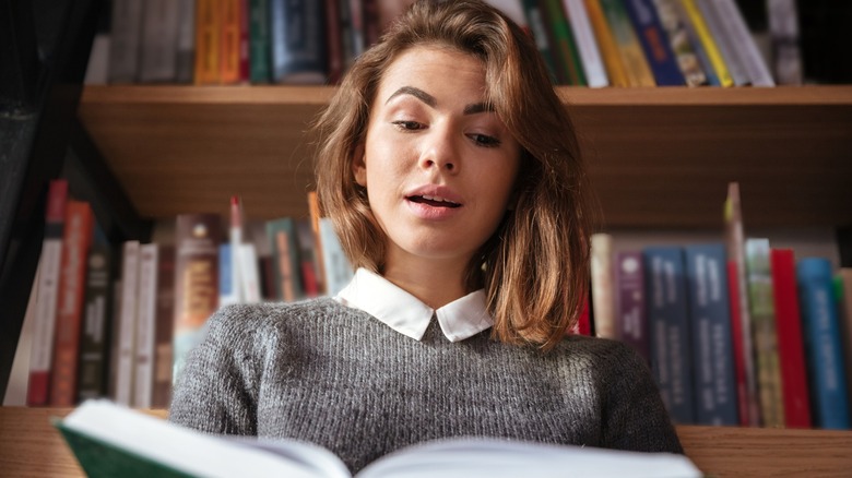A brunette woman reading a book while sitting in front of a bookshelf
