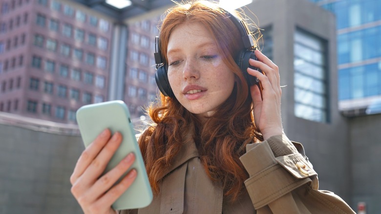 A woman with red hair wearing over ear headphones and looking at her phone while outside