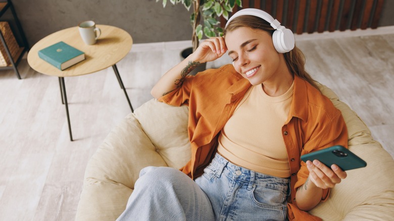 A woman with her eyes closed sitting in a chat while wearing headphones and holding her phone
