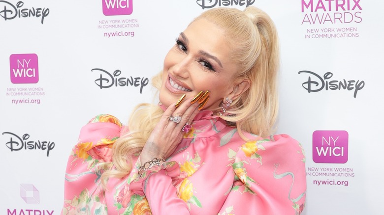 Gwen Stefani in a pink outfit smiling with her hand on her cheek on the red carpet