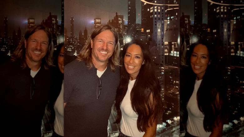 Joanna Gaines posing with her husband Chip in front of a window showing a city skyline