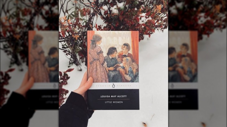 A hand holding up a copy of "Lilttle Women" with cut branches in the background