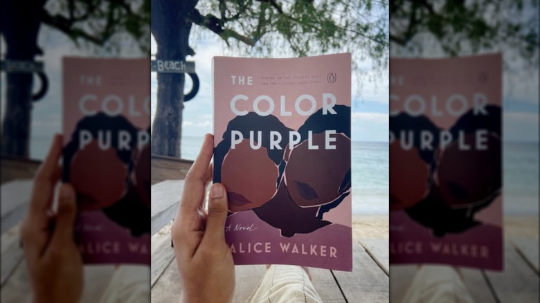 A hand holding up a copy of "The Color Purple" with a body of water in the background