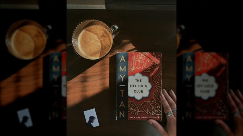 A copy of "The Joy Luck Club" on a table next to a cup of coffee and a hand with rings