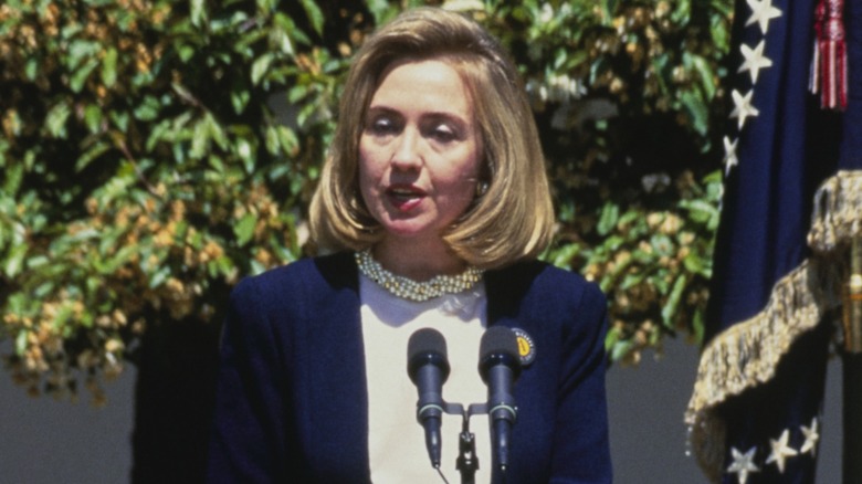 Hillary Clinton speaking into a microphone on a podium while outdoors in 1995
