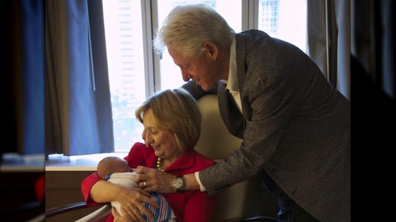 Hillary Clinton sitting in a chair holding her granddaughter Charlotte while Bill Clinton leans in
