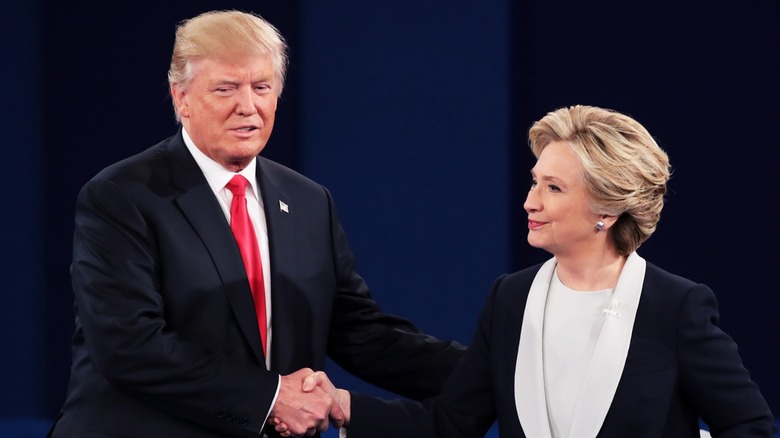 Donald Trump shaking hands with Hillary Clinton in front of a dark blue background in 2016