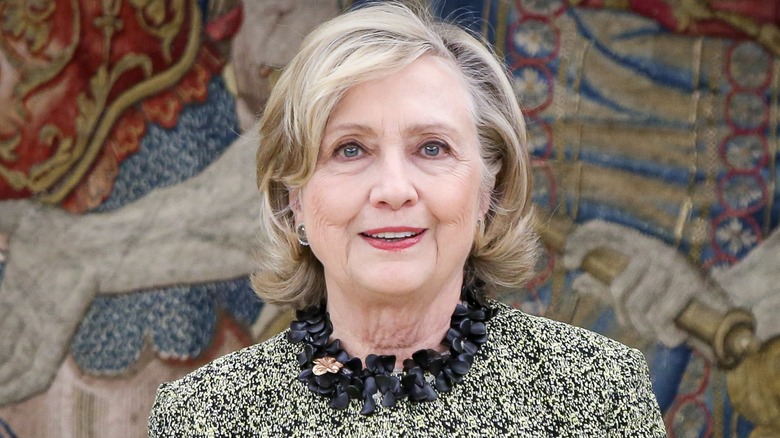Hillary Clinton slightly smiling in front of a mural wearing a statement necklace and patterned top