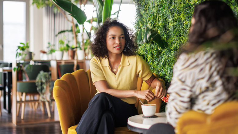 Two women sitting in chairs in a plant-filled cafe and engaged in conversation