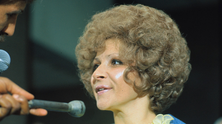 Brenda Lee speaks behind a microphone with a blonde voluminous perm