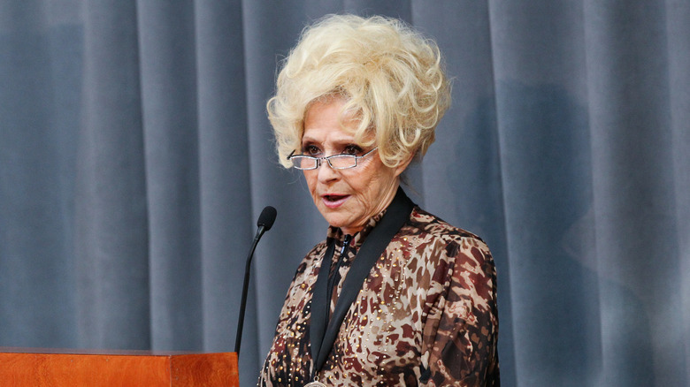 Brenda Lee speaks into a microphone wearing an animal-print blouse and glasses