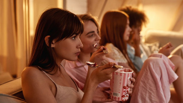 Group of four women, eating ice cream and wearing PJs while watching unseen TV