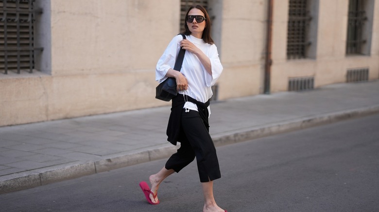 A woman wearing a white top and black capri pants while wearing red sandals and sunglasses