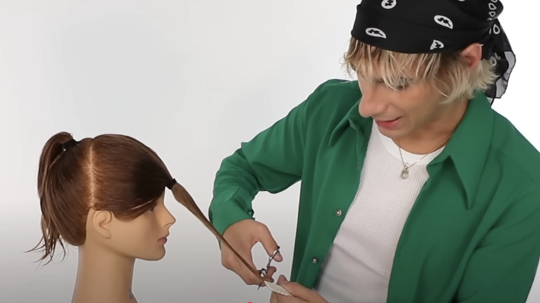 A man n a green shirt cutting hair on a mannequins head