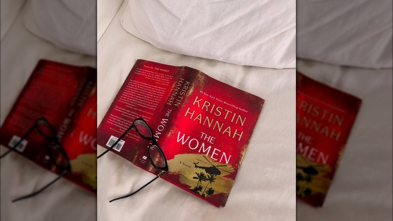A copy of "The Women" laying open on a bed with some reading glasses