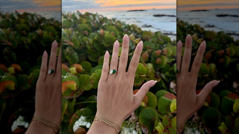 A hand with a diamond and emerald engagement ring with plants and ocean in the background