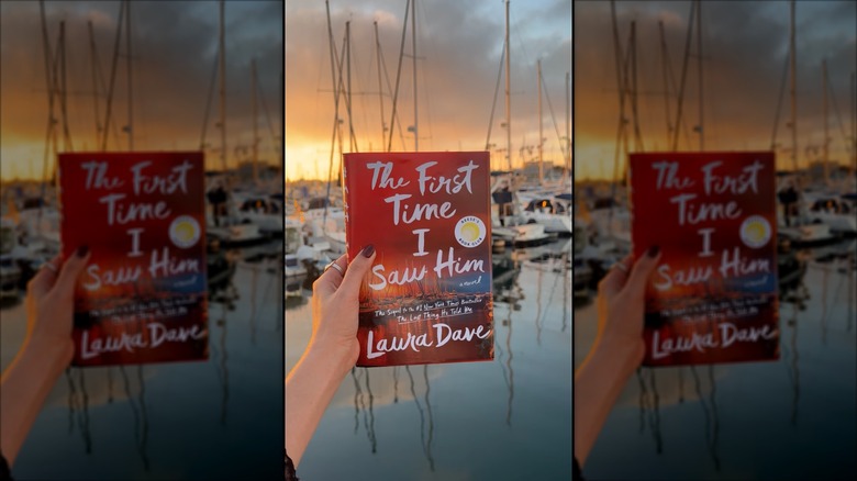 A hand holding a copy of "The First Time I Saw Him" up in front of a harbor full of sail boats